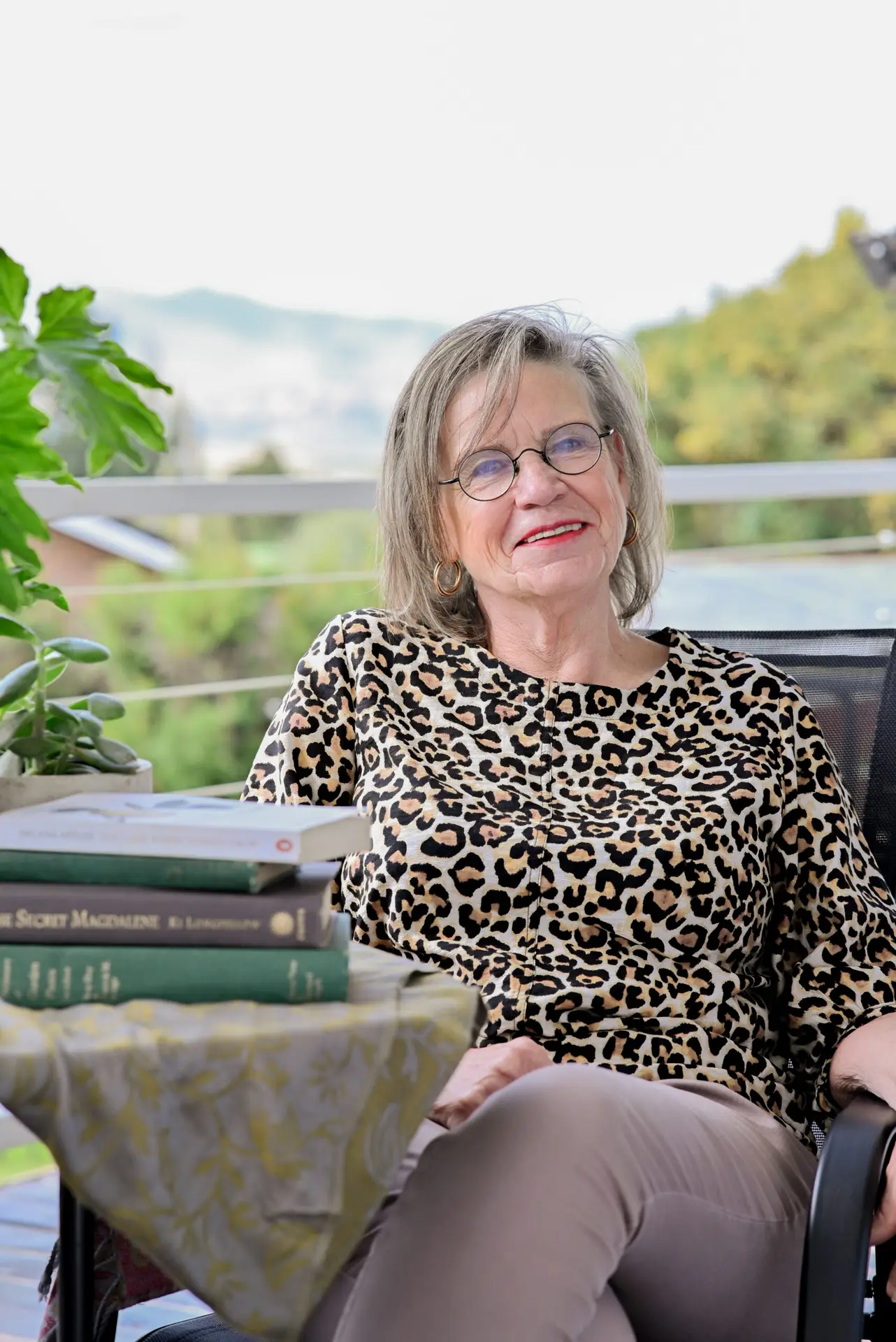 An older woman with glasses and a leopard-print top sits outdoors on a patio chair, smiling. There are books and a potted plant on a table beside her. Trees and mountains are visible in the background.