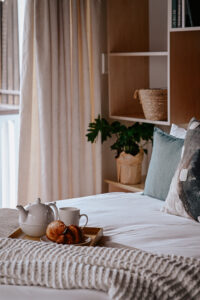 A cozy bedroom with a neatly made bed, a tray holding a teapot, cup, and croissants, soft pillows, a textured blanket, and plants near a window with light curtains.